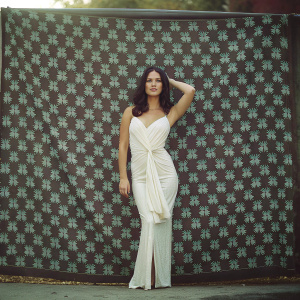 Olivia Munn in white dress with wrap design at award show. Large branded wallpaper background.