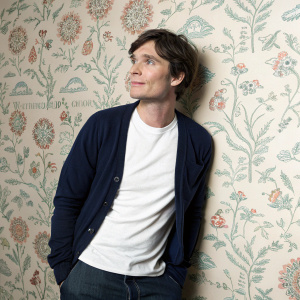 Actor Cillian Murphy in a casual outfit. Wearing a white t-shirt and navy cardigan. Against a busy wallpaper background.