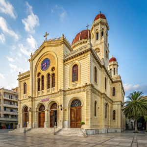 Photograph of Agios Minas Cathedral, Heraklion, Crete. Ornate cathedral with light stone exterior. Large arched entrance, numerous arched windows with colorful stained glass. Set against an urban backdrop.