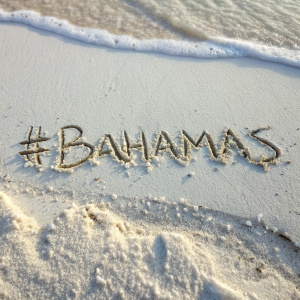A macro photo of the hashtag #Bahamas etched into white sand. Beach scene with grains still shifting.