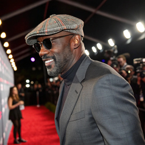 Idris Elba at the red carpet of the Film Independent Spirit Awards. Gray suit, plaid cap, and sunglasses. Glowing lights and paparazzi in the background.