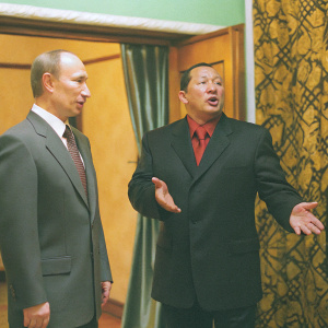Close-up candid shot of Vladimir Putin and Hugo Chavez. Putin stands slightly to the left, Chavez to the right, both in dark suits. Chavez makes an open-handed gesture. Patterned wallpaper and a green curtain form the background.