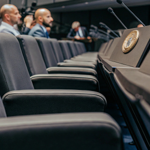 Pep Guardiola, sitting in a black chair at a press conference. Manchester City's badge prominently displayed on the table in front of him.