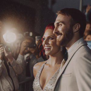 Sharna Burgess and James Hinchcliffe, smiling for photographers on a bustling blue carpet event.