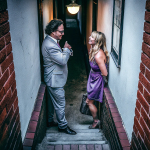 Johnny Galecki and Kaley Cuoco from 'The Big Bang Theory'. Leonard in gray suit, Penny in purple dress. Brick hallway with lamp. They are chatting.