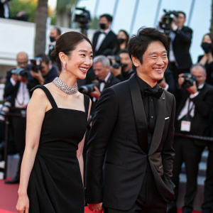 At the Cannes Film Festival, Jung Ryeo-Won and Kim Ki-Bang are walking the red carpet. Ryeo-Won in a chic black dress, Ki-Bang in a tailored suit. Both smile brightly for photographers.