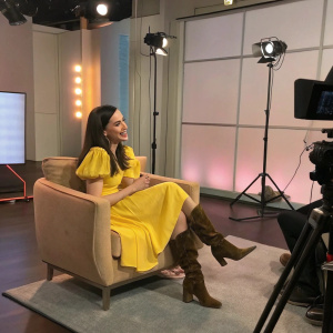 Sofia Carson in a casual interview setting. Wearing a vibrant yellow dress and brown boots. Chat show studio background.