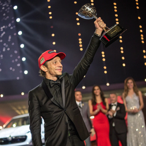 Valentino Rossi accepting an award at a motorsports gala. Wearing a sleek black suit and red cap, holding a trophy aloft.