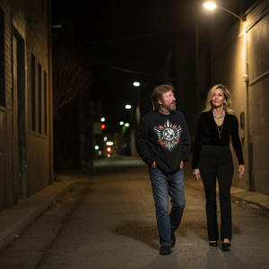 Chuck Norris and Gena O'Kelley strolling down a dimly lit alleyway. Chuck dons a black sweatshirt with a graphic design and casual attire. Gena is elegantly dressed in a sleek black outfit.