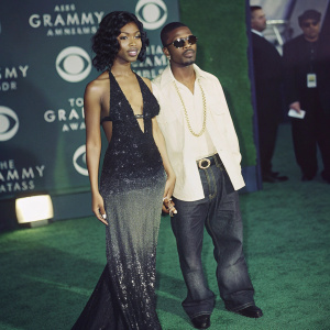 Brandy Norwood and Ray J on the Grammy red carpet. Brandy in a black and gray sequined gown, Ray J in a white shirt and sunglasses, gold chain necklace. The backdrop features Grammy logos and text.