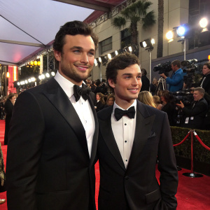 Tom Welling and Jamie White at a glamorous awards ceremony. Both are dressed in tuxedos. Crowded and lit red carpet.