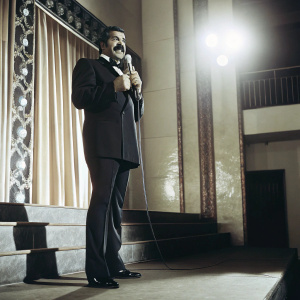 Shad Khan, with a prominent mustache, in a black suit, addressing a crowd. Jaguars logo and TIAA Bank signage visible on a bright, illuminated stage backdrop.