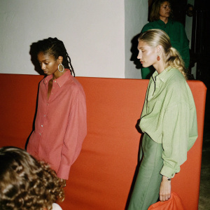 Chanel Iman and Elsa Hosk, models, standing at the center of a crowded fashion gala. They are wearing casual attire. Chanel Iman in a pink blouse, Elsa Hosk in a lime-green shirt. The background is a plain white wall.