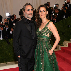 Joaquin Phoenix, in a sleek black suit, stands beside Rain Phoenix, elegantly dressed in a green gown with intricate detailing, at a gala event for Amazon Studios. Red carpet atmosphere, paparazzi flashes