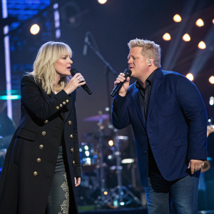 Natasha Bedingfield and Gary Levox, both in formal attire, perform a duet on an illuminated stage. Natasha, blonde hair cascading to her shoulders, wears a black coat with intricate button detailing. Gary, with silver hair, is dressed in a navy blue blazer. They hold microphones, illuminated by a variety of stage lights, against a backdrop of musical instruments and stage equipment.