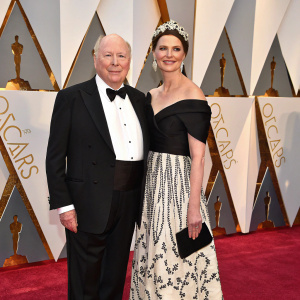 Julian Fellowes, in a classic black suit, and Emma Joy Kitchener-Fellowes, in an elegant black and white dress, posing on the red carpet. Golden statues and shiny awards all around.