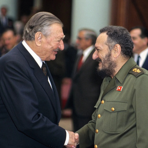 David Rockefeller and Fidel Castro formally shaking hands at a diplomatic conference. Attendees in formal wear, blurry background.