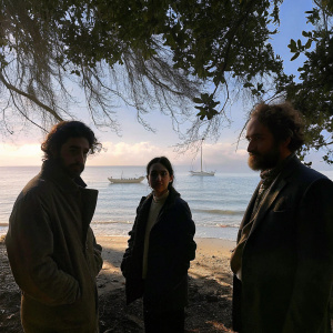 Golshifteh Farahani, Vincent Macaigne, and Louis Garrel on a serene beach. Clear blue sky, boats on the horizon. Natural lighting and candid pose.