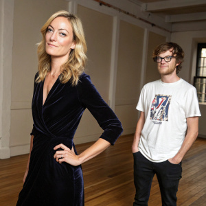 Lucy Walker, blonde hair, in black velvet dress. Kevin Pearce, casual attire, white t-shirt, graphic design. They pose indoors with wooden flooring, neutral background, relaxed stance.