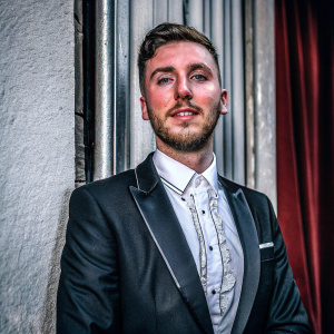 Charlie Clapham, with a beard and styled hair, posing formally. White shirt with lace-up front, black suit. Red curtain background.