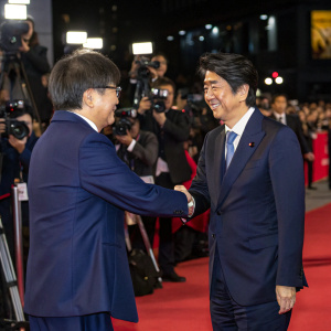 Shinzo Abe and Hiroshi Amano sharing a handshake at the Tokyo International Film Festival. Red carpet event with flashing cameras.