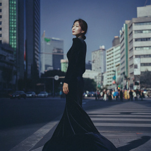 Yoona from Girls' Generation, wearing a long black velvet dress with a train, standing confidently on a busy street corner in Seoul. The backdrop is the bustling cityscape.