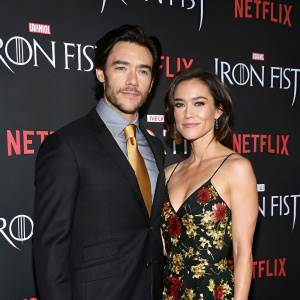 Tom Pelphrey and Jessica Stroup photographed at the 'Iron Fist' event in NYC. Tom in dark suit with gold tie, Jessica in floral gown. Background: Netflix and Marvel logos