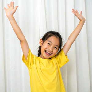 Close-up of a young Asian girl with a bright smile. She's wearing a yellow t-shirt, hands up, posing against a white curtain.