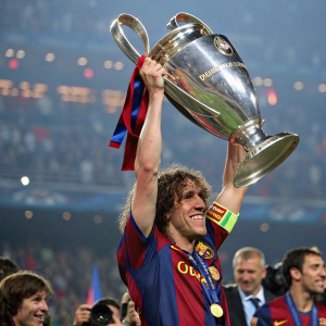Carles Puyol, curly-haired soccer star, victorious and holding aloft the UEFA Champions League trophy. He's dressed in Barcelona's iconic blue and red jersey. Surrounded by teammates and fans in a packed arena.