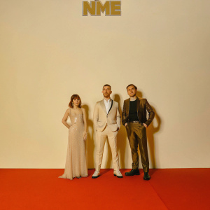 Formal group portrait at the NME Awards. Lauren Mayberry, Martin Doherty, Iain Cook stand against a black backdrop adorned with NME logo. Red carpet beneath their feet.