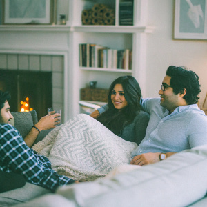 Kunal Nayyar, Neha Kapur, and Jason Kim gathered around a cozy fireplace in a living room setting. Warm ambient lighting