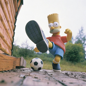 In a still frame from The Simpsons, Bart Simpson in mid-action, orange shirt, blue shorts, special sneakers, kicking a black and white soccer ball. Dark object near his foot, white background.