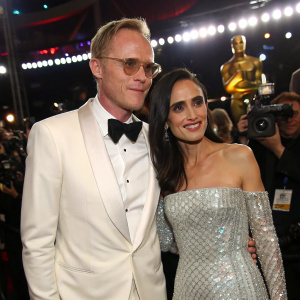 Paul Bettany, in a sharp white tuxedo, and Jennifer Connelly, in a stunning silver gown, celebrating at a bustling award show. A sea of cameras and flashing lights fill the background.