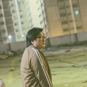 Close-up of Masi Oka in a gritty cityscape. Dark hair, glasses, serious expression. Wearing a brown jacket with white stripes.