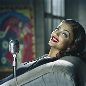 Aishwarya Rai Bachchan, close-up. Dark hair, makeup, red lipstick. Smiling confidently. Black top, relaxed pose. Microphone in front. Blurred colorful backdrop.