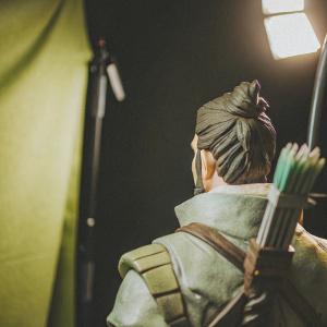 Sculpture of Overwatch character Okami Hanzo, showcasing his detailed back view. Made of textured gray and brown materials, holding a bow and quiver. Shot in a studio with focused lighting against a black backdrop.