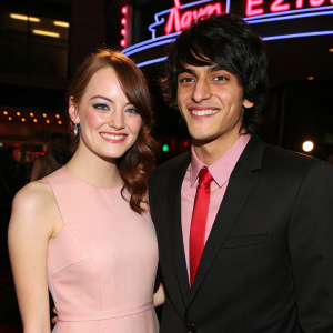 Emma Stone and Teddy Geiger at award show. Emma in a pastel pink dress, Teddy in black suit with red tie. Both smiling. Backlit neon sign