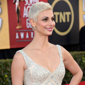 Morena Baccarin, with platinum blonde hair and a radiant smile, at a glitzy awards show. She is dressed in a stunning sleeveless, white gown adorned with shimmering patterns. The backdrop is filled with bold logos and graphics.