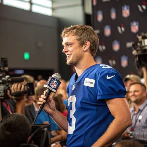 Jake Browning, wearing a blue NFL jersey, at the post-game press conference with a microphone in hand. A sea of cameras and journalists in the background.