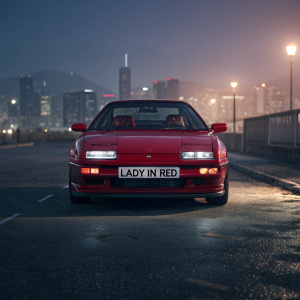 Illustration of a sleek red Honda Prelude. Parked on a dimly lit street. License plate reads 'LADY IN RED'. City skyline in the background.