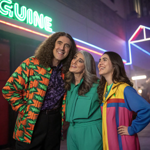 Weird Al Yankovic, Suzanne Yankovic, and Nina Yankovic posing for a music video shoot. Neon lights flicker in the background