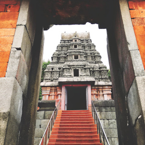 An awe-inspiring photograph showcasing the grand entrance of Prasanna Virupaksha Temple. The structure, built with layers of aged gray stone blocks, is intricately carved and features a central pathway flanked by a spiraling staircase.