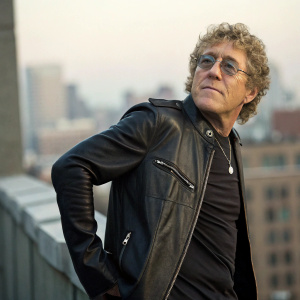 Roger Daltrey, curly hair, glasses, dressed in a leather jacket, posing against a blurred cityscape background.