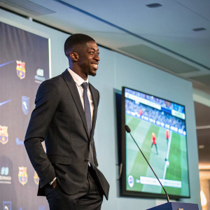 Ousmane Dembele at a press conference, wearing a suit. The backdrop features a large screen showcasing soccer highlights.
