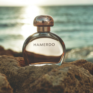 A luxury perfume bottle labeled 'HAMERDO' placed on textured coastal rocks, with a calm sea and soft sunset in the background. The lighting is natural and balanced, capturing subtle golden tones while maintaining a neutral and fresh atmosphere. The sea reflects the sky with hints of blue and gentle ripples, creating a tranquil and elegant scene, without overwhelming warm hues