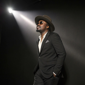 Anthony Hamilton at the 59th Grammy Awards. Wearing a black suit, hat, and sunglasses. Solid black background, spotlight effect