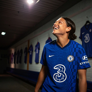 Sam Kerr, a vibrant and joyful soccer player, stands out in her Chelsea FC Women's jersey. She's inside a sports hall adorned with jerseys lining the walls. Dimly lit with a spotlight casting a radiant glow on her.