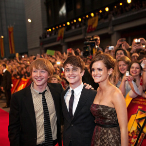 Rupert Grint, Daniel Radcliffe, and Emma Watson at a Harry Potter reunion event. Crowded venue with fans holding cameras and waving banners.