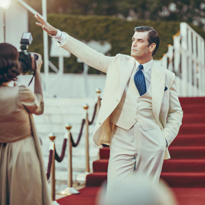 Pierce Brosnan in a sharp navy suit, crisp white shirt, and royal blue tie, striking a pose at a glamorous awards show. Bright studio lighting, no background