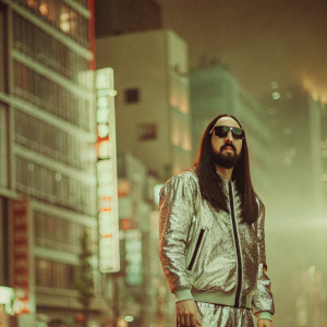Steve Aoki, wearing a futuristic silver jacket and dark sunglasses, standing in front of a neon-lit cityscape.